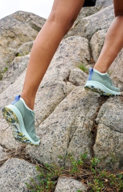 Light green trail running shoes with patterned soles climbing over large gray rocks in an outdoor setting.