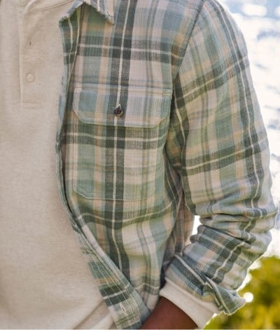 Person in green plaid shirt over beige henley stands outdoors with sunlight on water behind.