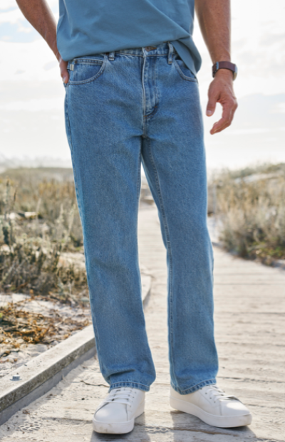 A man wearing blue jeans, a blue shirt, and white sneakers stands on a wooden path in an outdoor setting.