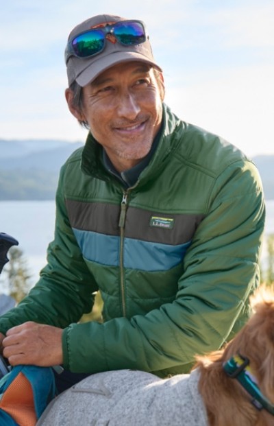 A man wearing a green jacket with blue and gray stripes, and sitting outdoors near a body of water.