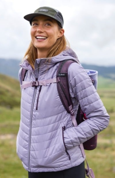 A woman in purple quilted jacket, cap, and backpack stands outdoors with green hills in the background.