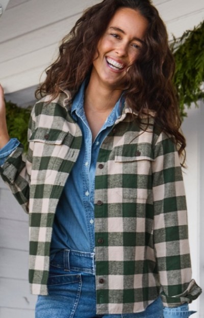Woman in green and beige checkered jacket over denim shirt stands in front of a white wall with greenery in the background.