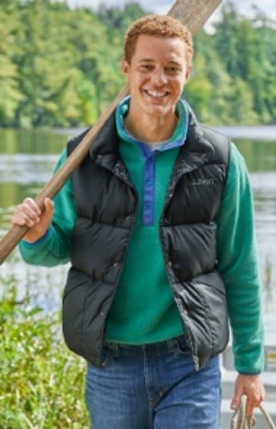 A man in a green sweater and black puffy vest, stands near a lake with trees in the background.