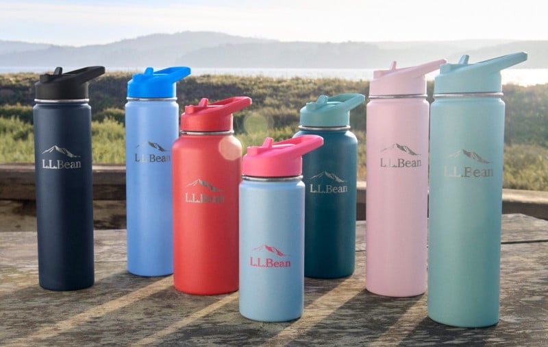 Seven colourful L.L.Bean water bottles of various sizes stand on a wooden table outdoors with a coastal background.