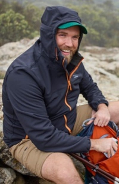 A man in outdoor gear, sitting on a rock, wearing a hooded jacket and shorts, holding an orange backpack.