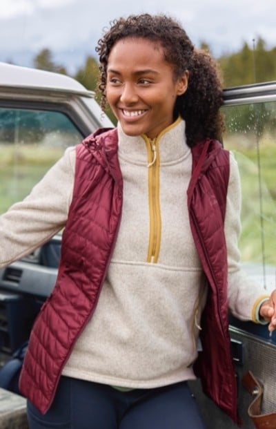 Person in maroon quilted vest over beige fleece with yellow zipper stands beside an open car door in an outdoor setting.
