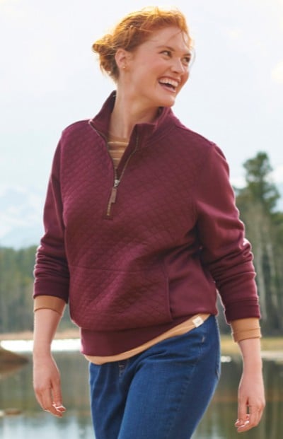 A woman in a maroon sweatshirt and blue jeans stands near water. The scene is outdoors and serene. 