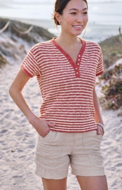 A woman wearing a red and white striped shirt with beige shorts stands on a sandy path leading to the beach.