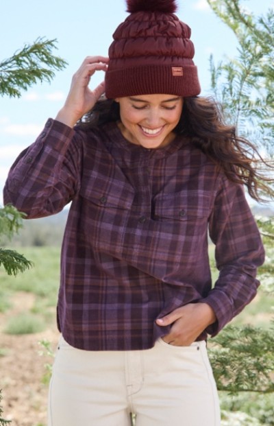 Woman in maroon beanie and plaid shirt stands outdoors among green trees, smiling and looking down. 