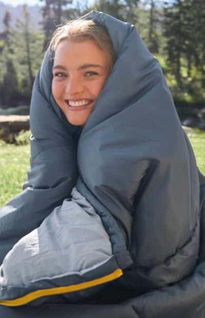 A woman wrapped in a gray sleeping bag, sitting outdoors on grass with trees and mountains in the background.