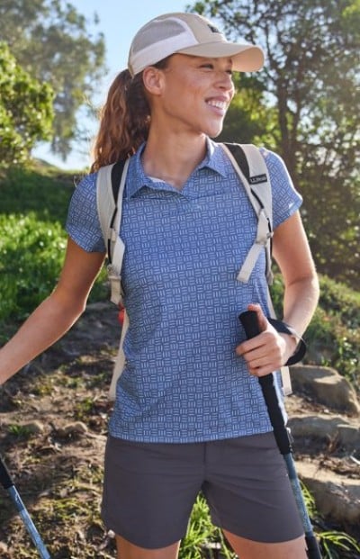 Wearing a cap, backpack, and hiking gear, the person holds trekking poles on a trail surrounded by greenery.
