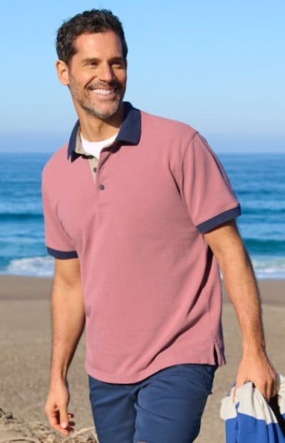 Person wearing a dusty rose polo shirt with navy trim and dark shorts while walking on a sandy beach.