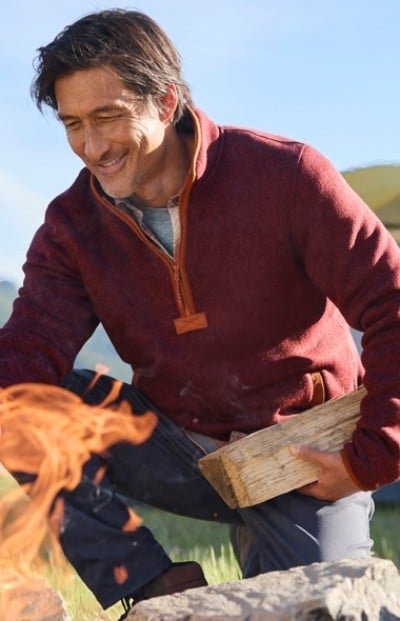 A person in a red sweater kneeling by a campfire, holding a piece of firewood.