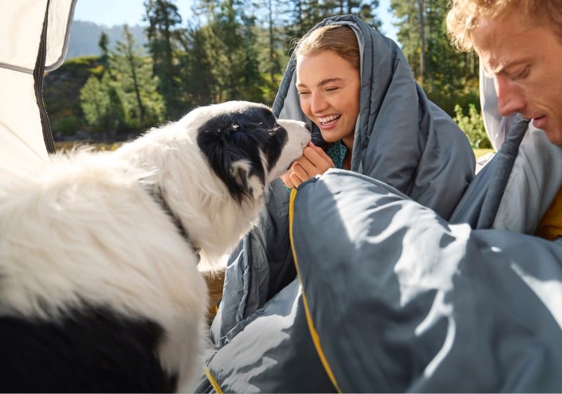 Man and woman wrapped in sleeping bags, sitting on the ground outside with a dog.