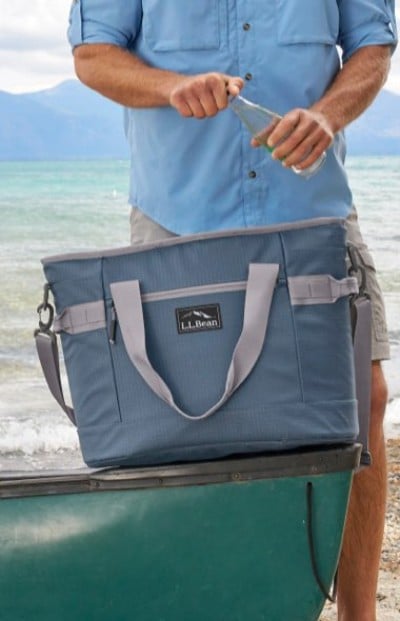 Blue L. L. Bean cooler bag rests on a green canoe while a person in a light blue shirt opens a bottle by the lakeshore.