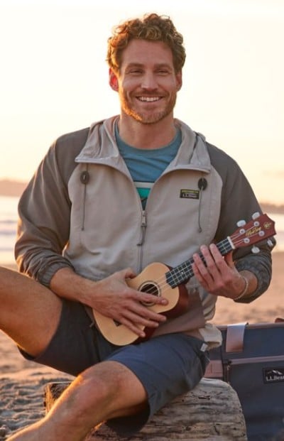 Person wearing a windbreaker sits on a beach at sunset, playing a small ukulele with a bag beside them.