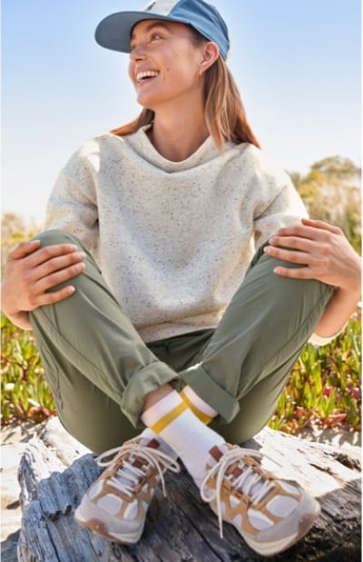 A woman wearing beige sneakers, a grey sweater and green pants sits cross-legged on a rock with hands on knees.