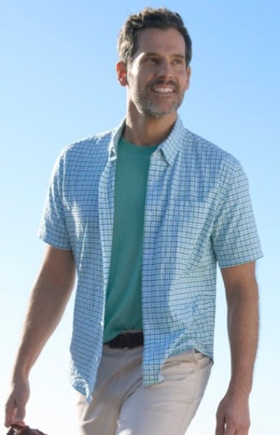 Person wearing a teal T‑shirt with a light blue short‑sleeve checkered shirt and light pants, walking outdoors under a clear sky.