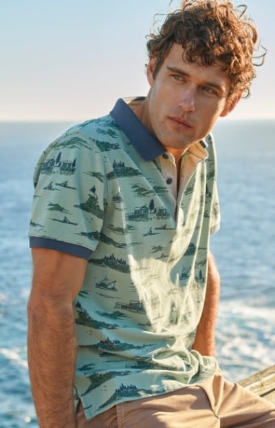 A man wearing a light green polo shirt with blue patterns and beige pants is sitting near the ocean