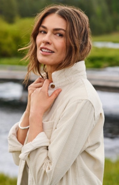 Woman outdoors in light-colored jacket stands near a river with greenery in the background. 