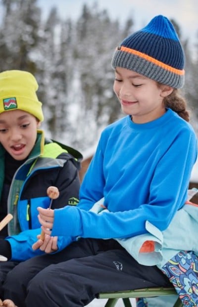 Two kids dressed for cold weather sit outdoors. One wears a blue sweater and beanie, the other a yellow hat and jacket.