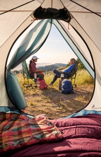 View from inside a tent toward campers seated in chairs at a campsite with a scenic mountain landscape in the distance.