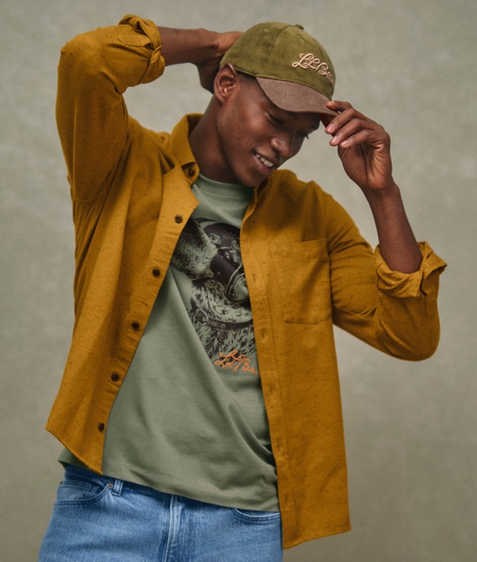 A man adjusting olive baseball cap, wearing an unbuttoned, mustard-colored shirt over a gray graphic tee.
