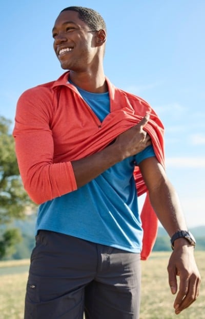 A man wearing a blue shirt and gray pants is putting on or taking off a red jacket outdoors on a sunny day.