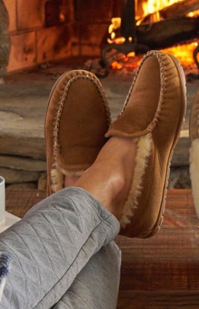 Cozy scene with tan shearling-lined slippers resting on a wooden surface near a stone fireplace with a warm fire burning.