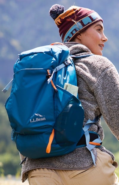 A person with a blue L L Bean backpack and a beanie hikes a trail outdoors, with mountains in the background.