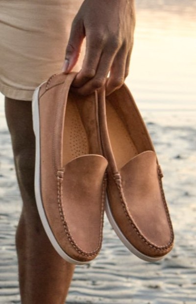 Close up image of a hand holding a pair of brown slipā€‘on shoes while standing on a sandy beach.