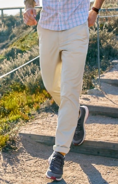 A man in beige pants and dark sneakers walks down outdoor wooden steps surrounded by greenery.