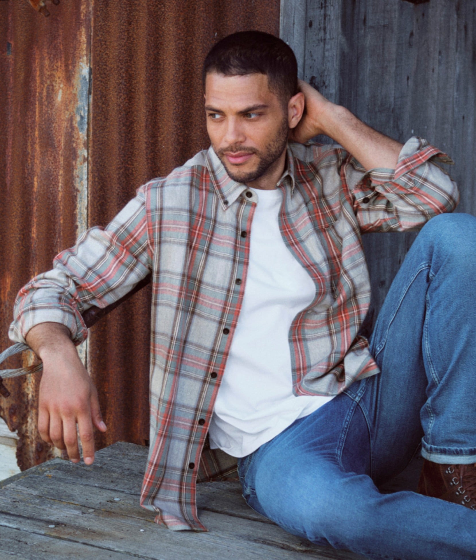 Man seated outdoors wearing a plaid flannel shirt over a white tee. 