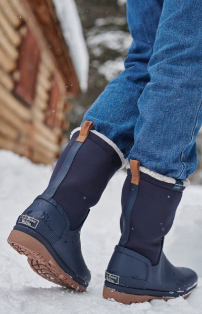 A woman in blue boots stands on snow, with a wooden structure in the background, capturing a moment of winter adventure.