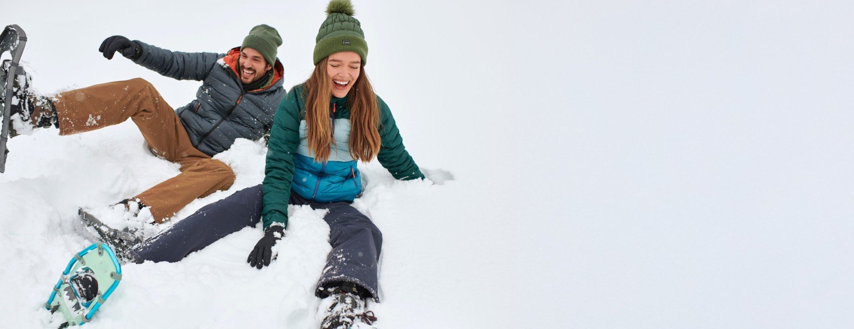 Two people in winter gear sit in deep snow with snowshoes on, appearing playful and relaxed outdoors.