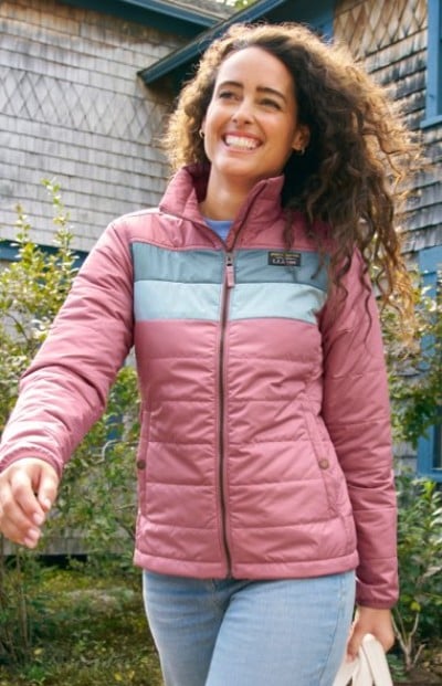 A person in a pink zip-up jacket with teal chest accents, styled for an outdoor casual look near a shingled building.