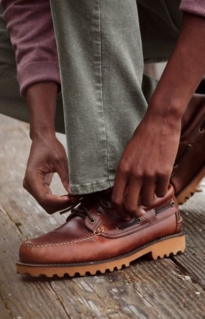 Close up of hands tying brown leather shoes while seated on a wooden deck.