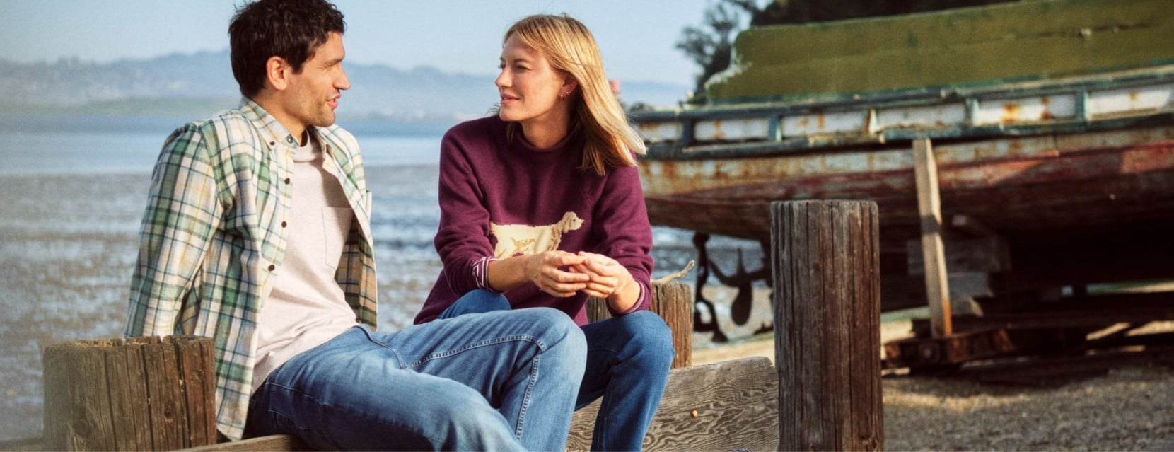 Two people sit on a wooden dock by the water, talking beside a weathered boat on a calm coastal shoreline.