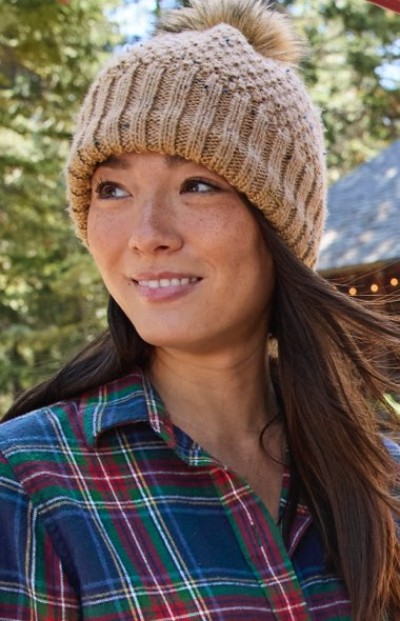Person in beige knit hat with pom-pom and plaid shirt stands outdoors with trees and a building in the background.