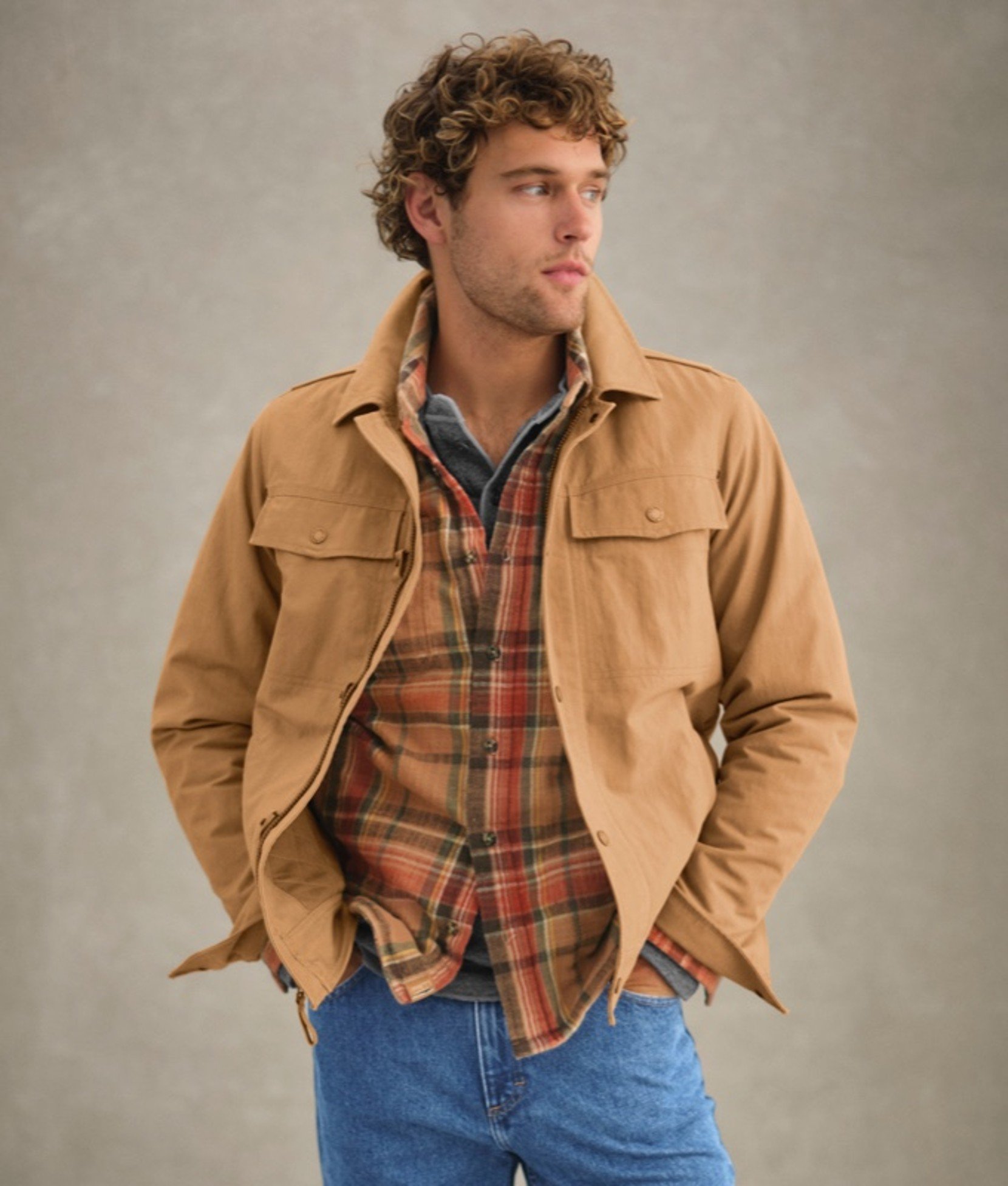 Man in tan jacket over plaid shirt with hands in pockets. 