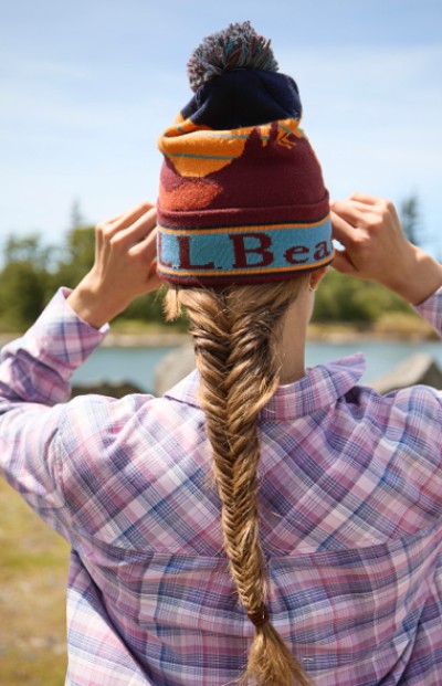 A woman is adjusting a beanie on their head outdoors, showcasing a long braided hairstyle.