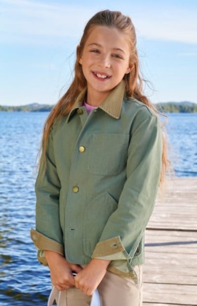 A child in a light green jacket with a tan collar stands on a dock near the water.