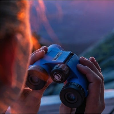 Close-up of someone holding binoculars, the setting sun casting orange-pink light on their face.