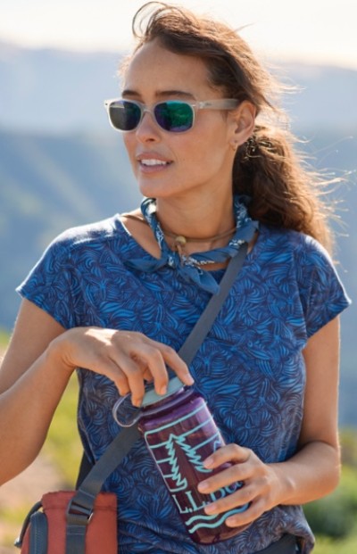 A woman in a blue patterned shirt holding a purple water bottle with a tree design, standing outdoors.
