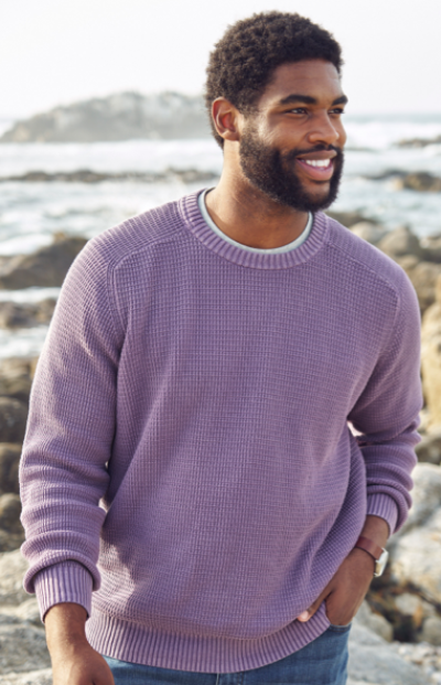 A man wearing a lavender sweater and jeans stands near a rocky shoreline with the ocean in the background.