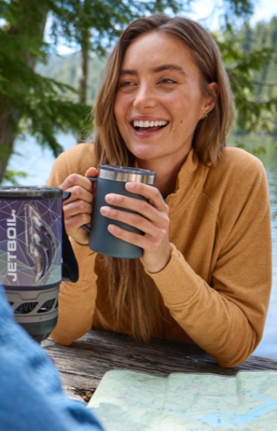 A woman sats at a picnic table, holding a mug, with a map and helmet on the table, set against a natural backdrop.