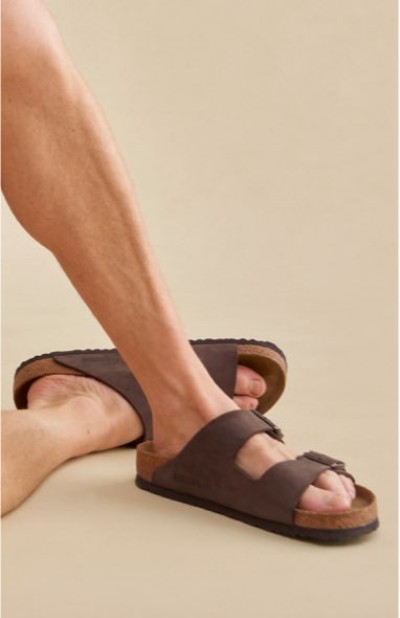 Close up of feet wearing brown leather slide sandals against a light studio backdrop.