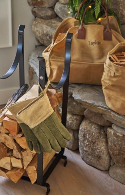 A fireplace set with gloves, wood, and a personalized bag rests by a stone hearth, creating a cozy and prepared atmosphere for a warm fire.