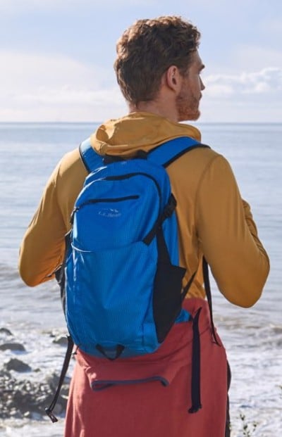 Person wearing a blue backpack and outdoor clothing, standing on a rocky shoreline overlooking the ocean.