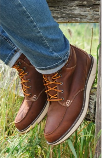 Close up of brown leather boots with yellow laces resting on a wooden fence above green grass.